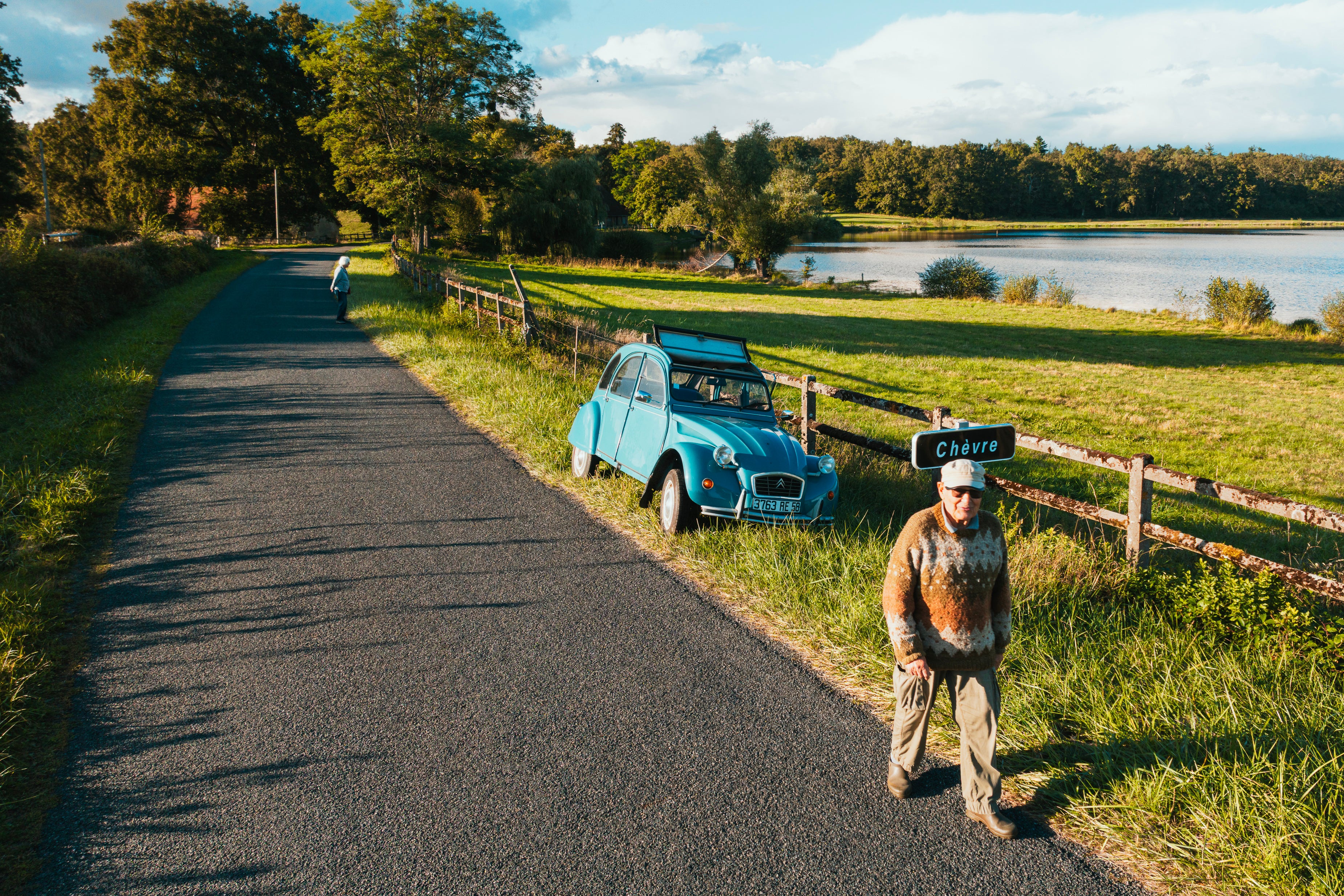 A blue Citroen 2CV parked on a country road beside a lake in the French countryside, my grandfather standing next to it.