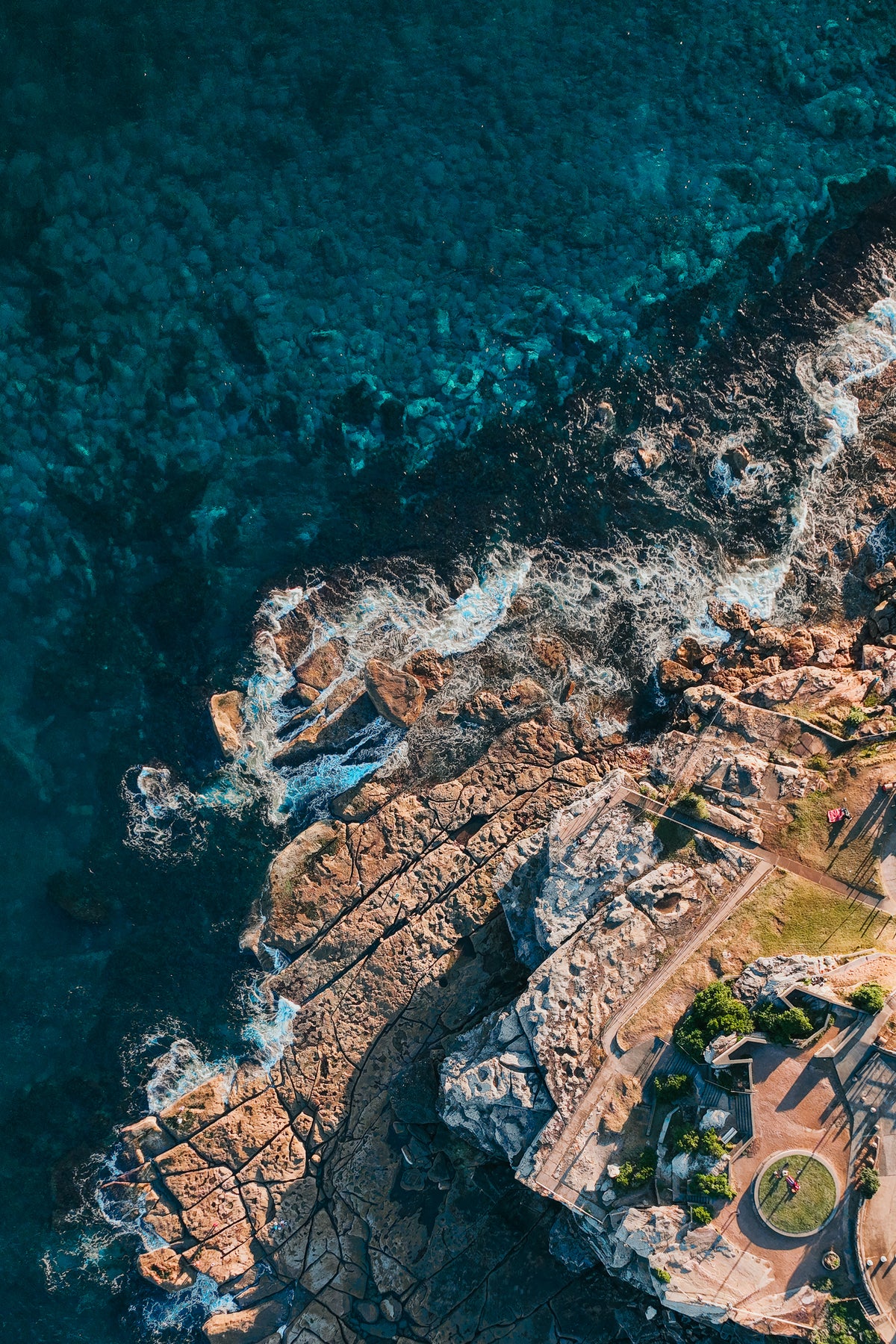 An aerial top-down view of deep blue ocean water crashing against a rugged sandstone cliff and a coastal walking path.