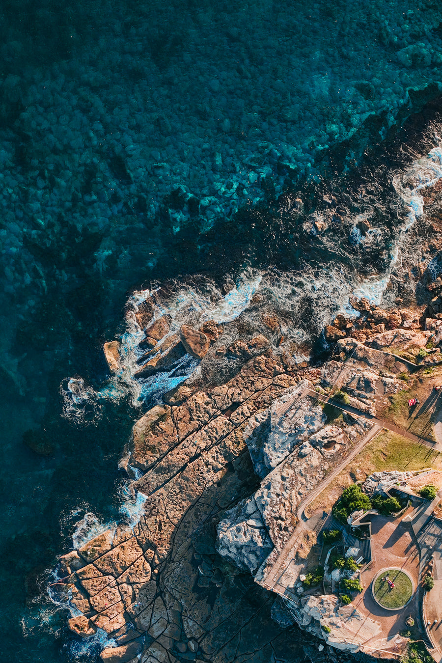 An aerial top-down view of deep blue ocean water crashing against a rugged sandstone cliff and a coastal walking path.