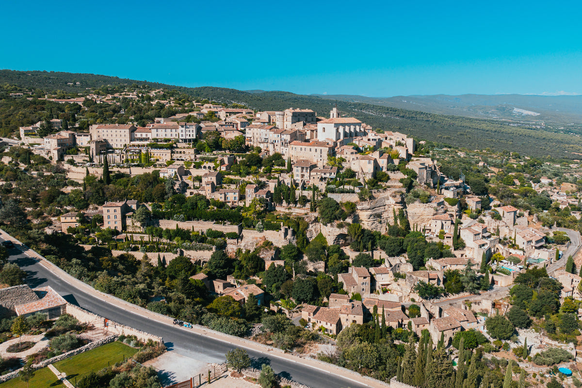 An aerial view of the historic stone village of Gordes nestled on a green hillside in Provence, France under a clear blue sky.