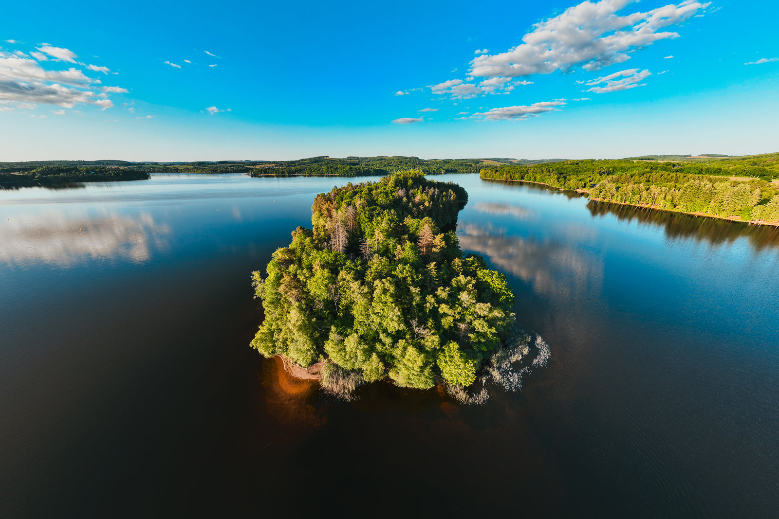 An aerial photograph of a lush green island in the center of a calm blue lake with cloud reflections.