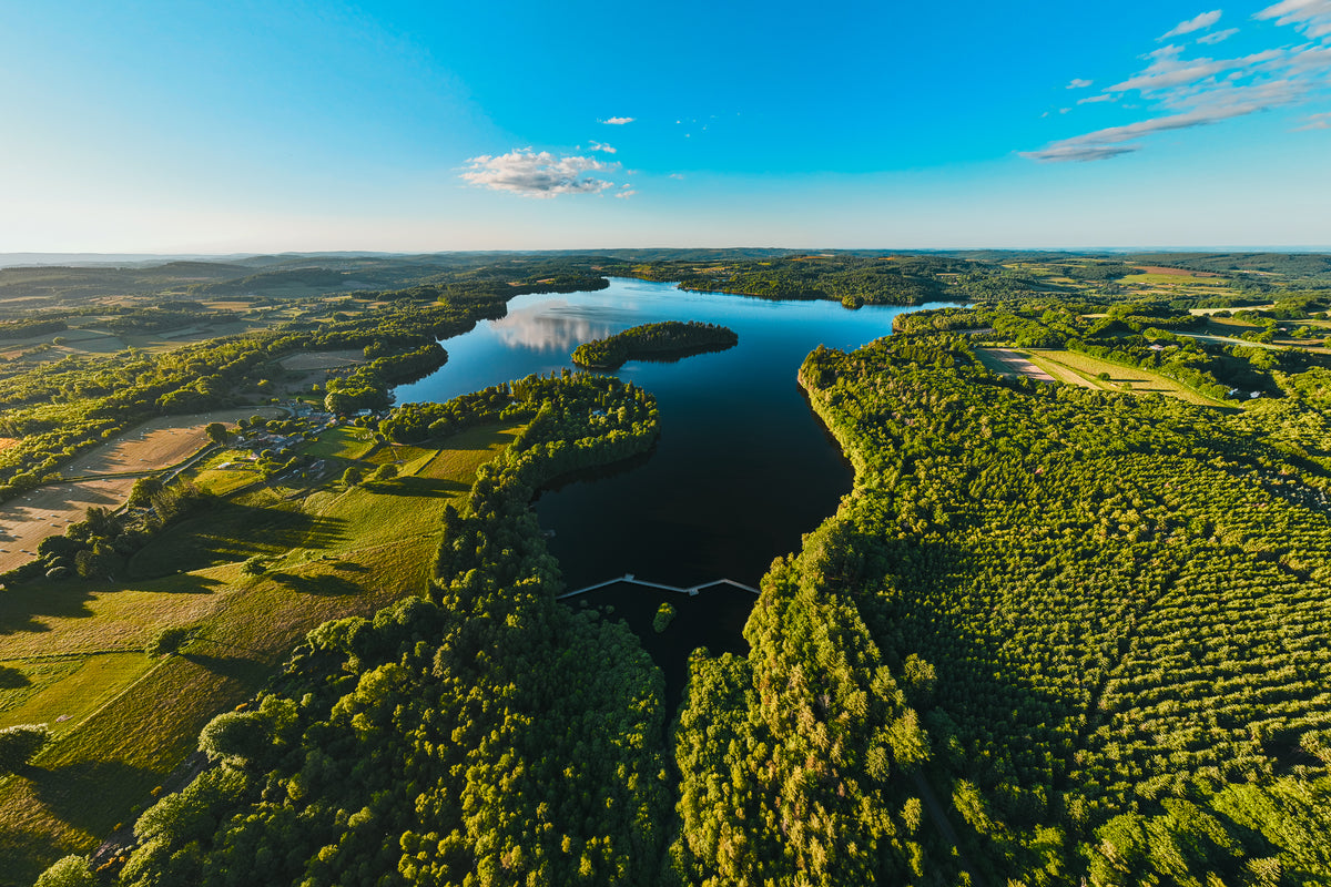 A wide aerial view shows a large blue lake with a small island surrounded by a dense green forest and rural fields under a clear sky.