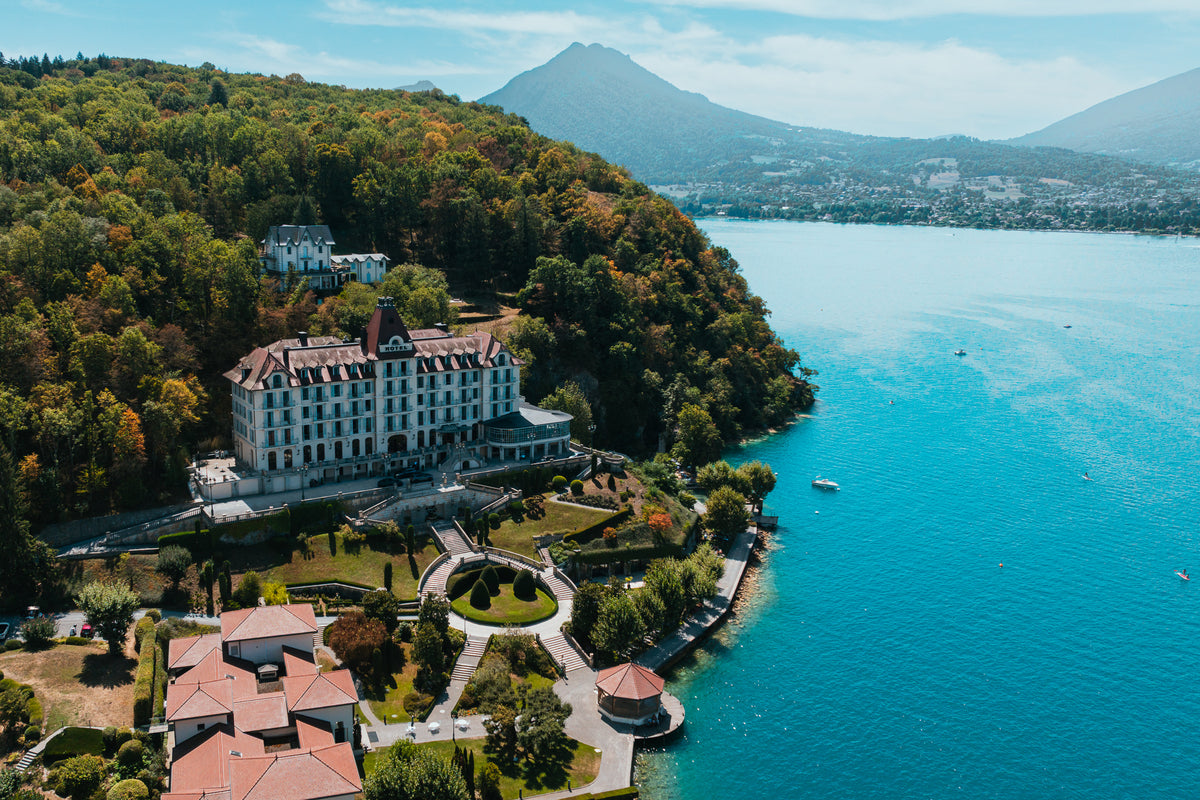 An aerial view of the grand Palace de Menthon hotel overlooking the bright blue waters of Lake Annecy in France.