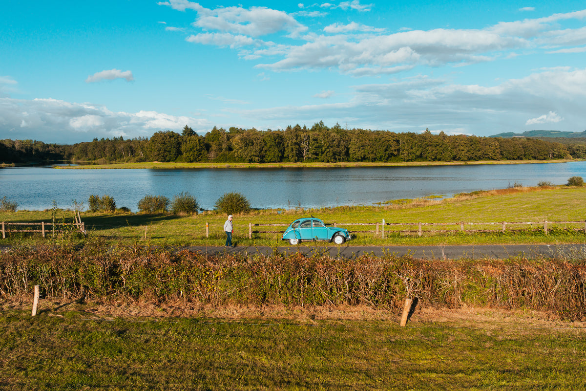 A vintage blue Citroën 2CV car parked on a rural road beside a lake in the French countryside.