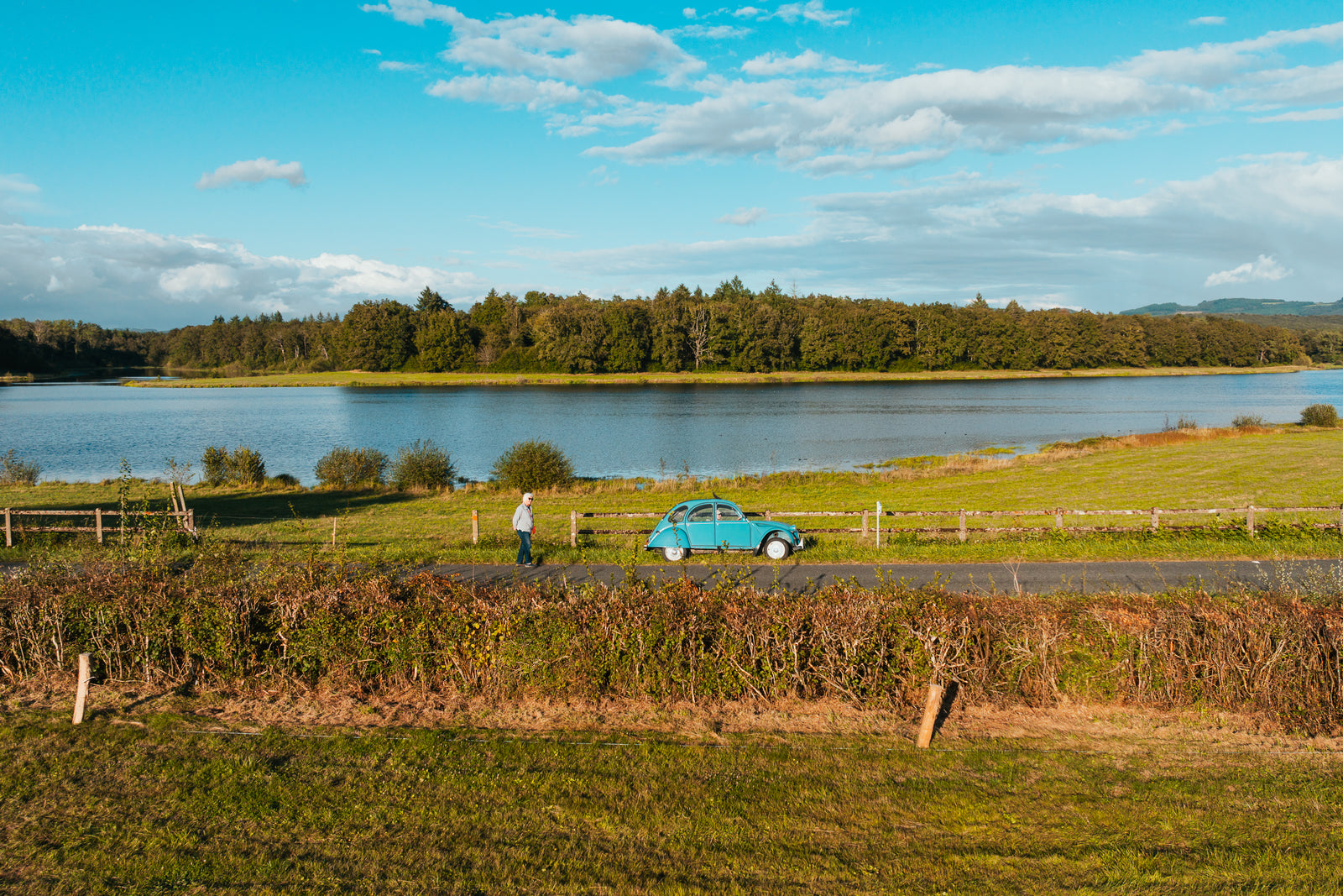 A vintage blue Citroën 2CV car parked on a rural road beside a lake in the French countryside.