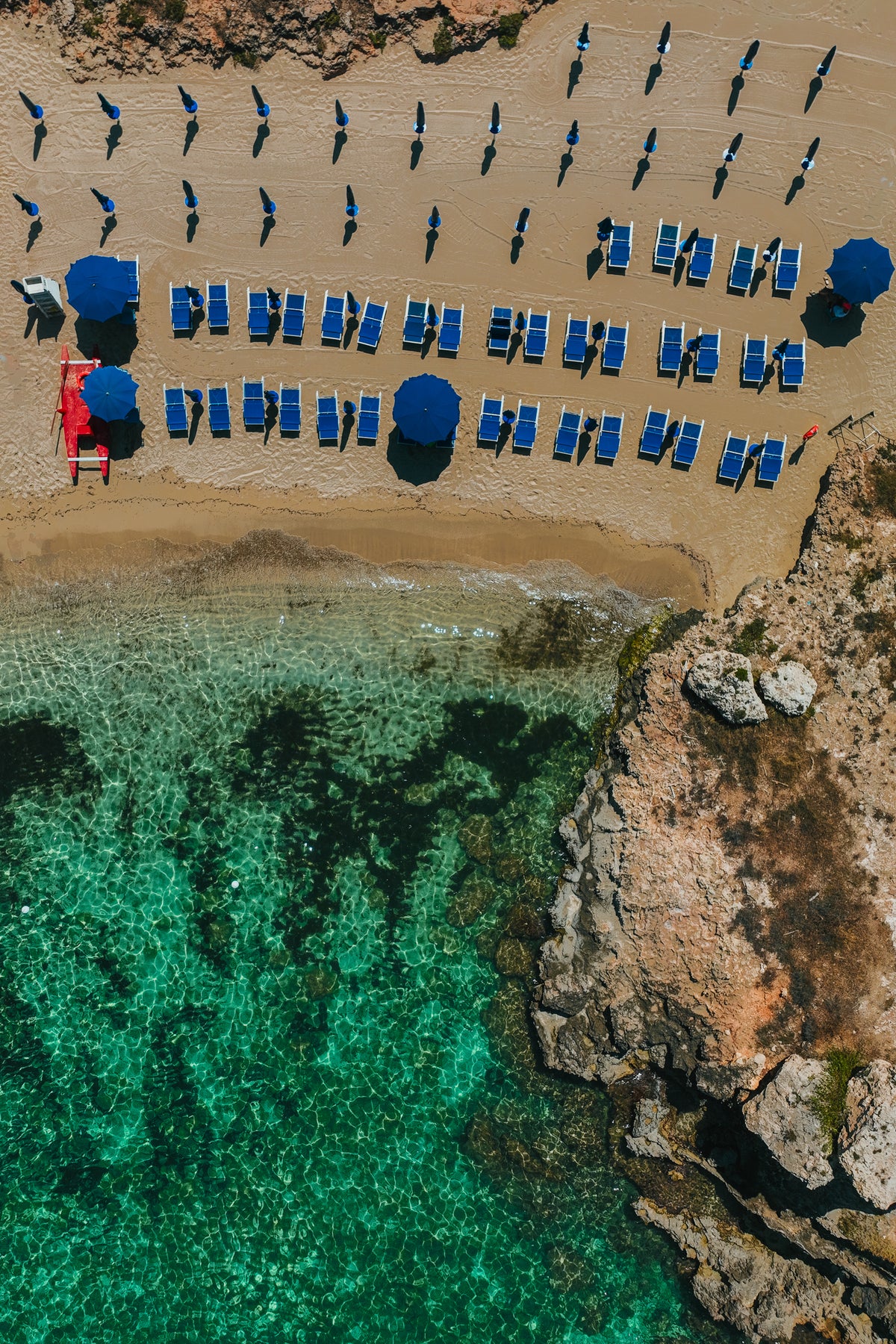 An aerial top-down photograph of a sandy beach in Italy with neat rows of blue sun loungers and umbrellas next to clear turquoise water and rocks.