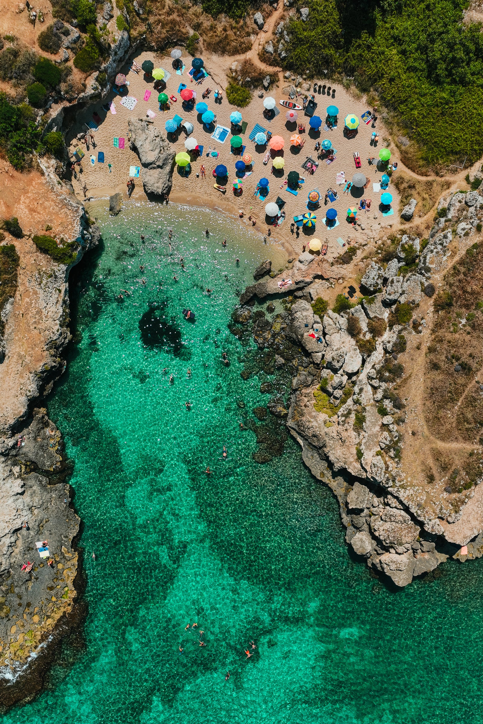 Aerial view of a small rocky beach cove in Italy with colorful umbrellas and people swimming in bright turquoise water.
