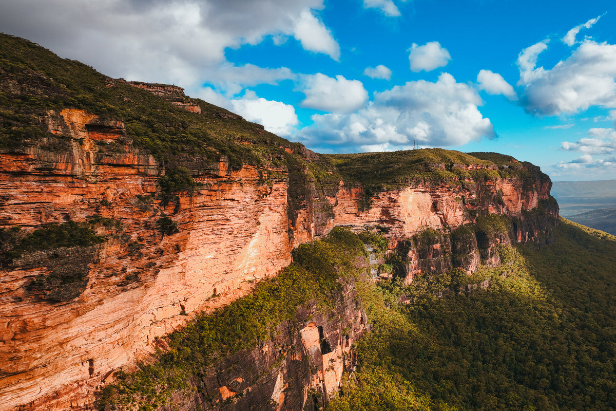 Sunlit red sandstone cliffs overlooking a vast green valley in the Australian Blue Mountains under a blue sky.