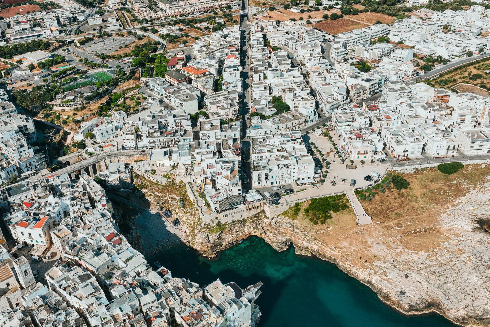 An overhead aerial view of the coastal town of Polignano a Mare in Italy, showing white buildings clustered on rocky cliffs above a turquoise sea cove.