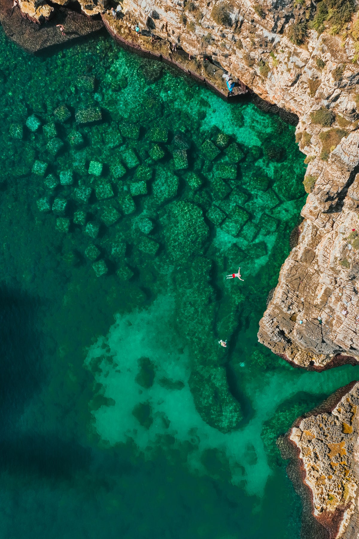 An aerial drone view looking down at two people swimming in the clear emerald and turquoise water next to rocky cliffs in Italy.