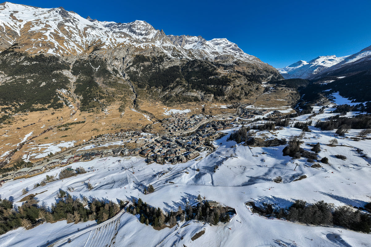 High-angle aerial photograph of the stone buildings of Val d'Isère village nestled in a valley between snow-covered ski slopes and rugged peaks.