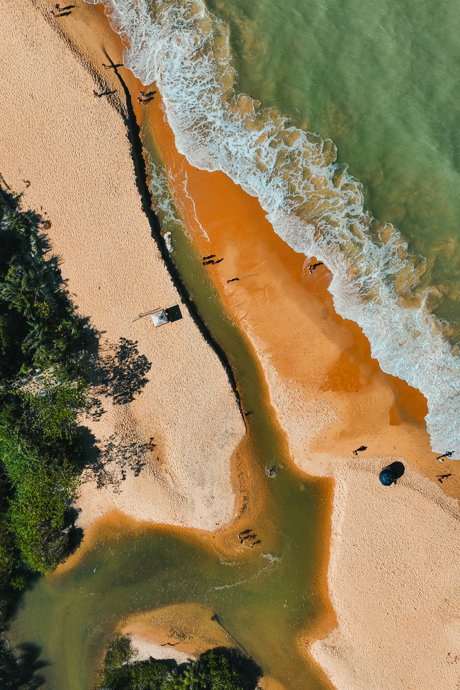 An aerial top-down view shows a narrow green river cutting through golden sand to meet the white foam of ocean waves in Brazil.