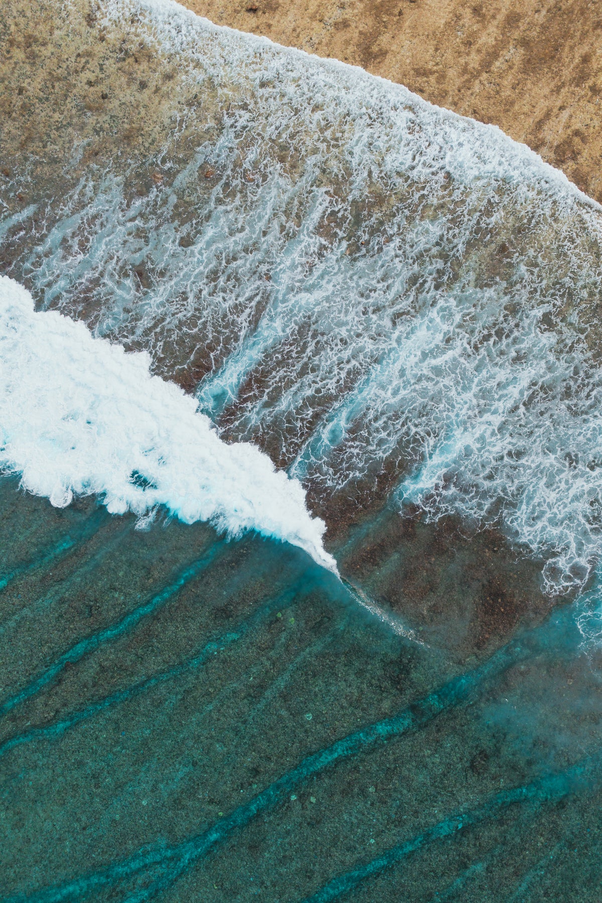 A vertical aerial view of a white foamy wave breaking over a dark reef into clear turquoise water next to a sandy beach.