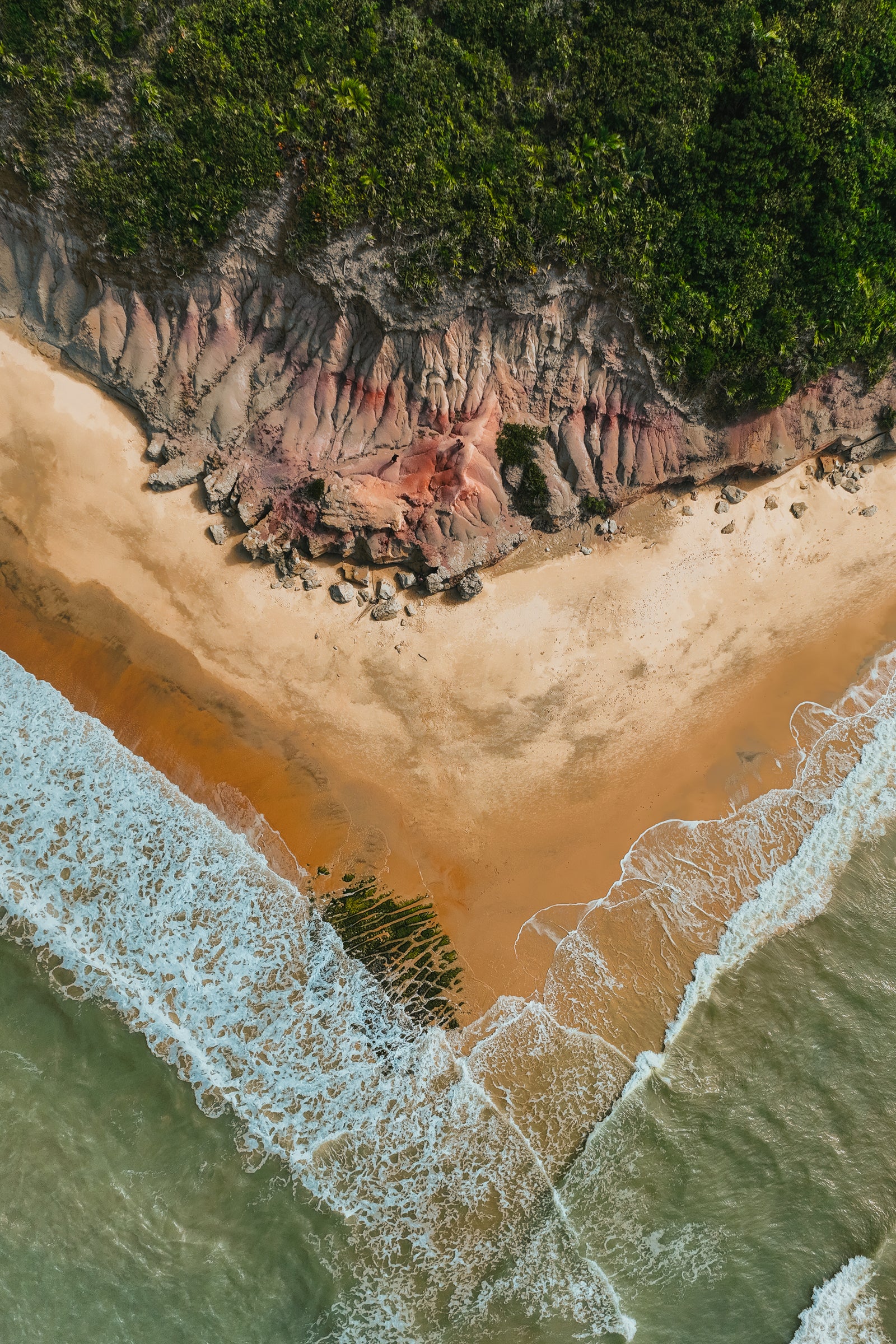 Top-down aerial view of a sandy beach where waves meet red textured cliffs and lush green tropical forest.