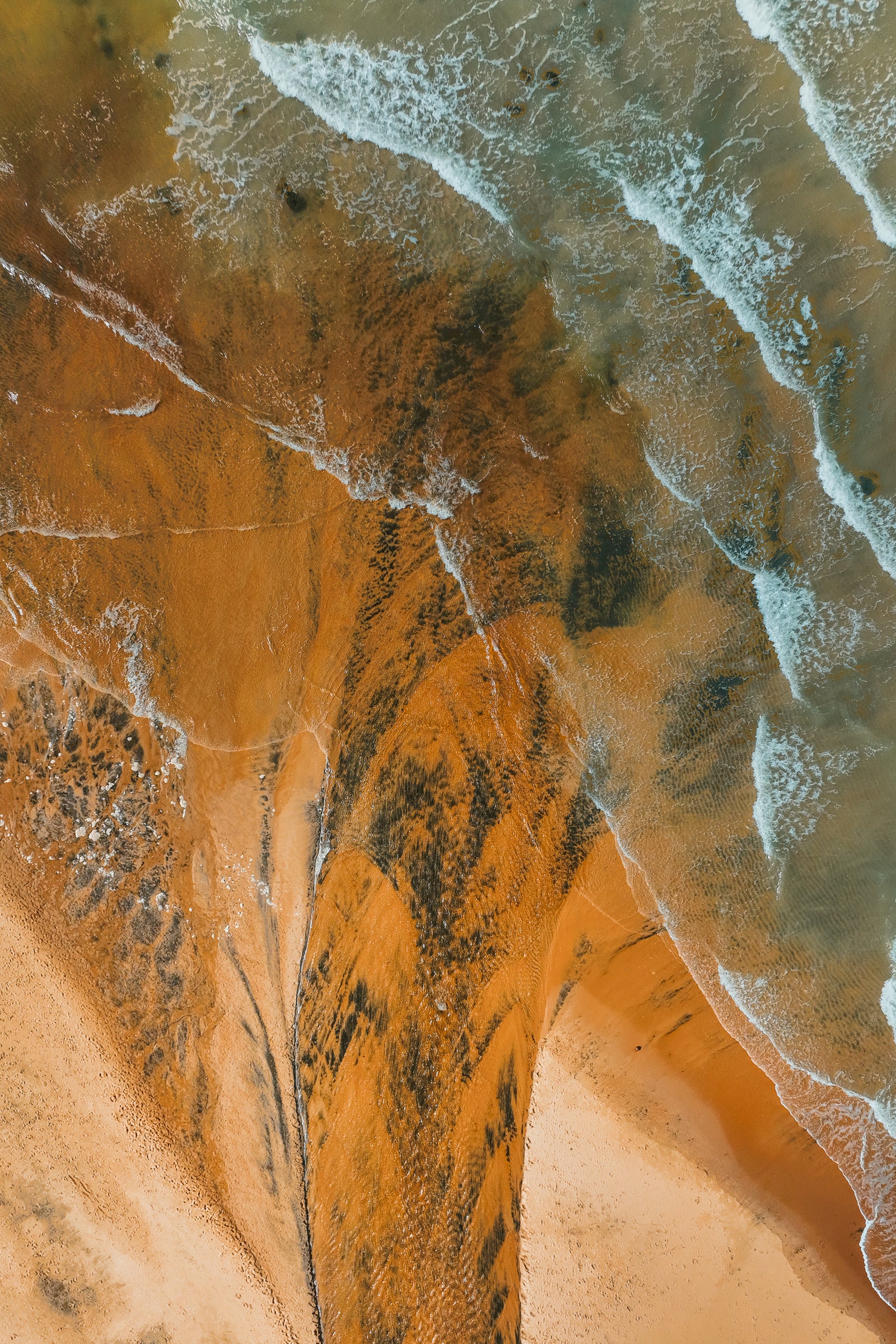 An overhead aerial photograph of a beach in Australia featuring striking orange sand with dark mineral patterns meeting gentle ocean waves.