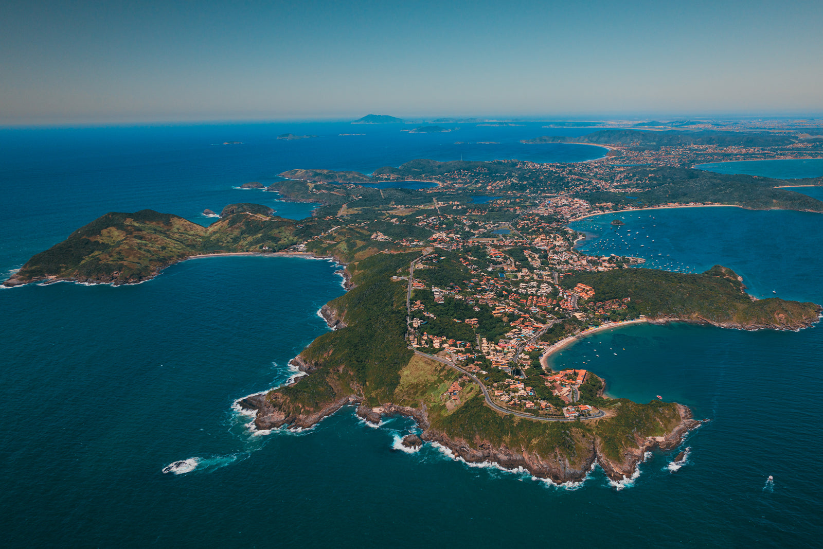 High-altitude aerial view of the lush green Búzios peninsula in Brazil jutting out into deep blue ocean waters under a clear sky.