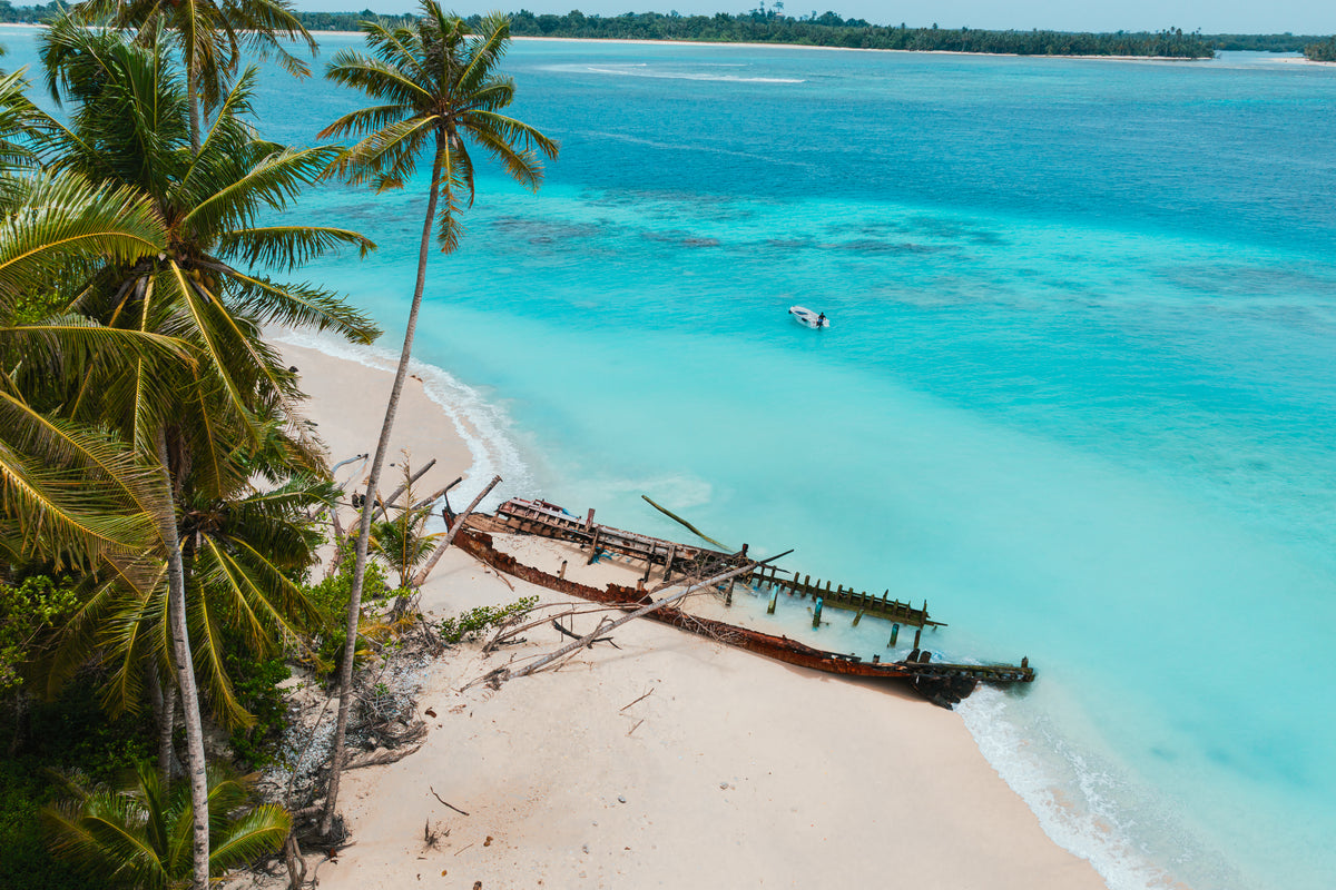 An aerial view of a rusted shipwreck resting on a white sand beach lined with palm trees and turquoise ocean water.