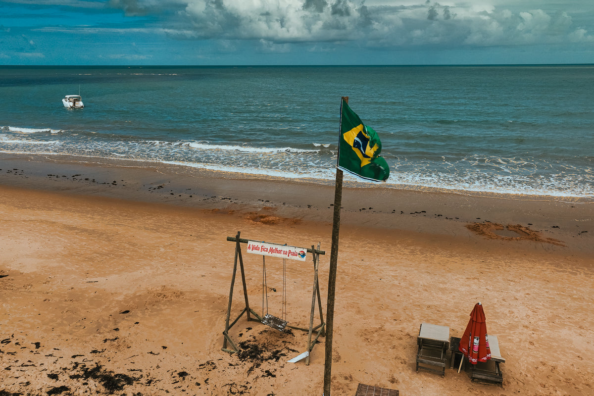 A Brazilian flag waving on a sandy beach with a small boat in the ocean and a wooden swing in the foreground.