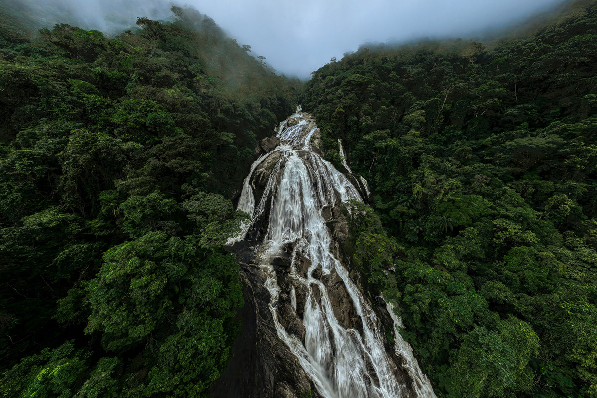 High angle view of a powerful waterfall cascading through a dense green rainforest under a cloudy sky.