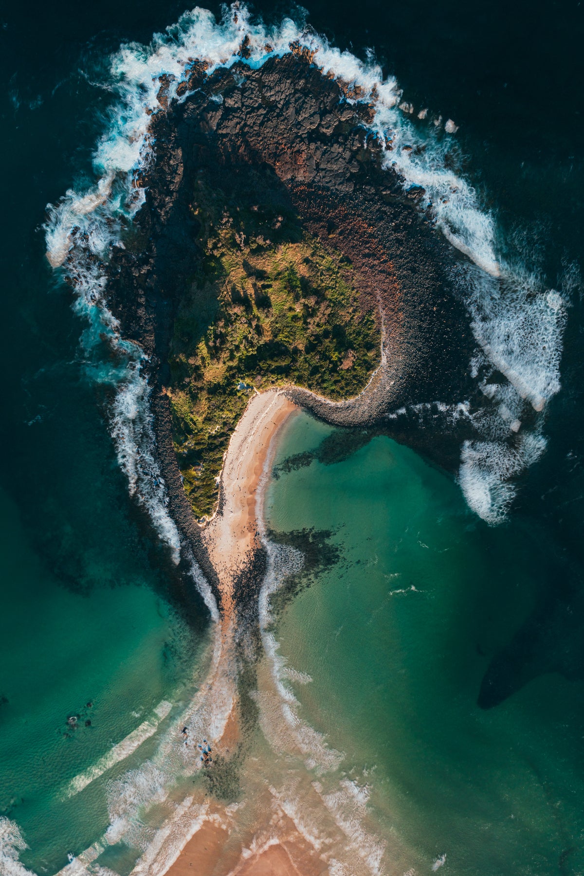 A vertical top-down aerial view of a rocky island connected to a sandy beach by a narrow spit of sand surrounded by turquoise water.