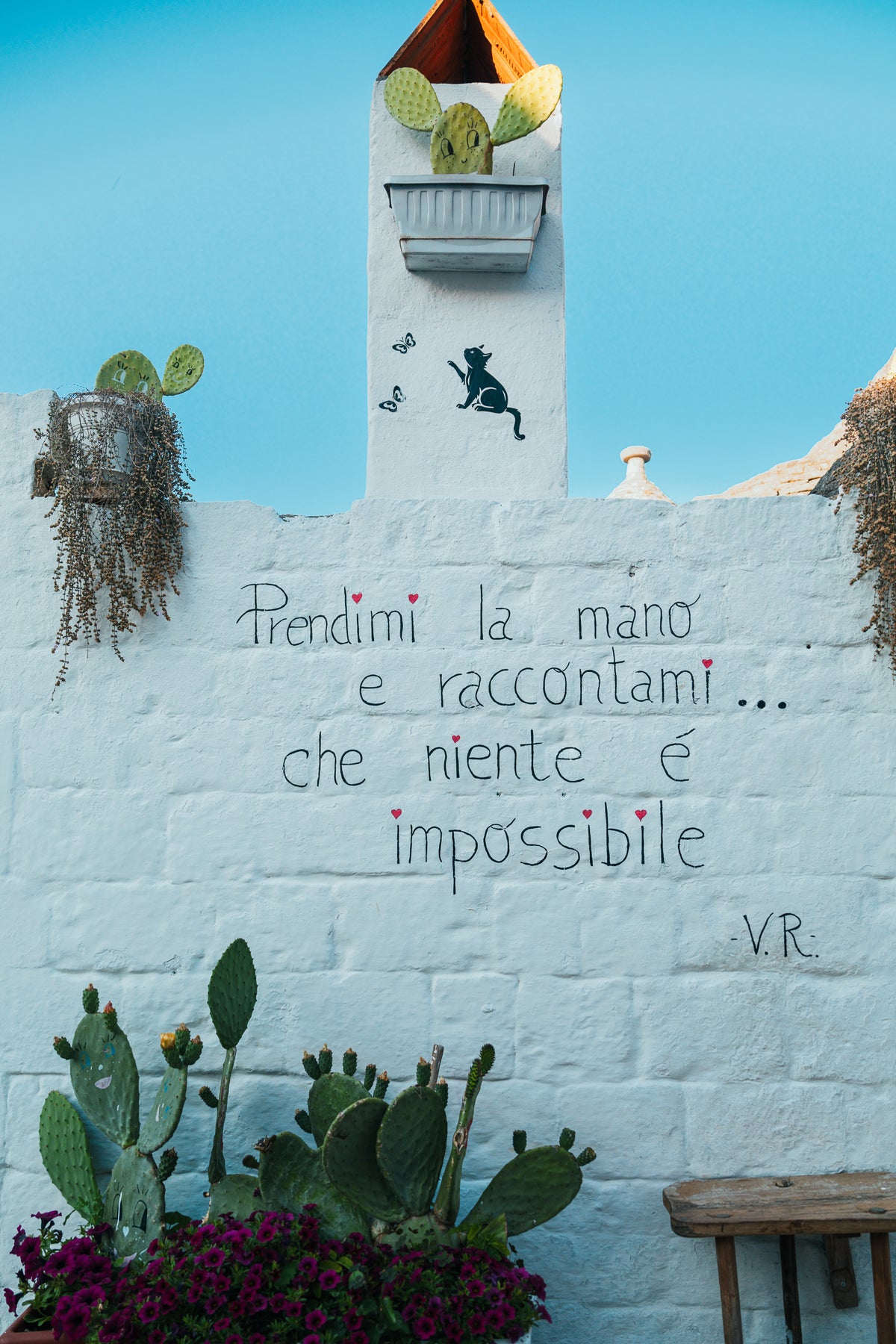 A white stone wall in Puglia with handwritten Italian poetry, prickly pear cacti, and a cat stencil against a clear blue sky.