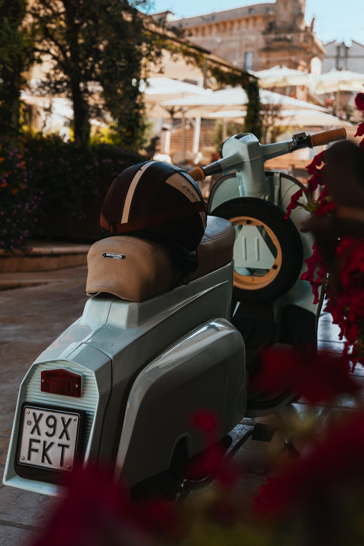 A vintage light blue Lambretta scooter parked in a sunny Italian square with a leather helmet on the seat and red flowers in the foreground.