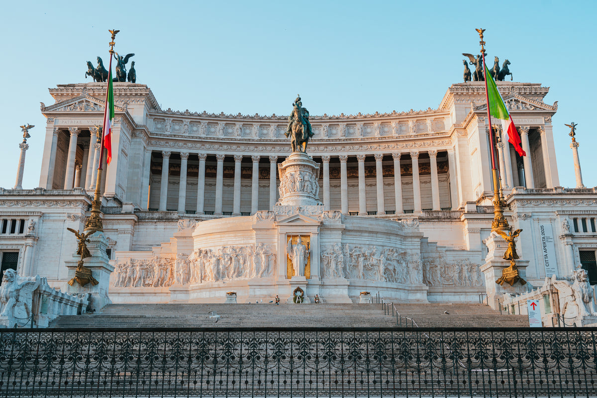 The white marble Altare della Patria monument in Rome stands against a clear blue sky with Italian flags on either side.
