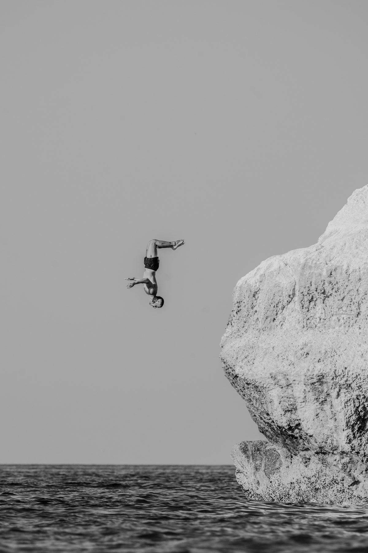 A minimalist black and white photograph of a man performing a backflip off a large white rock into the sea.