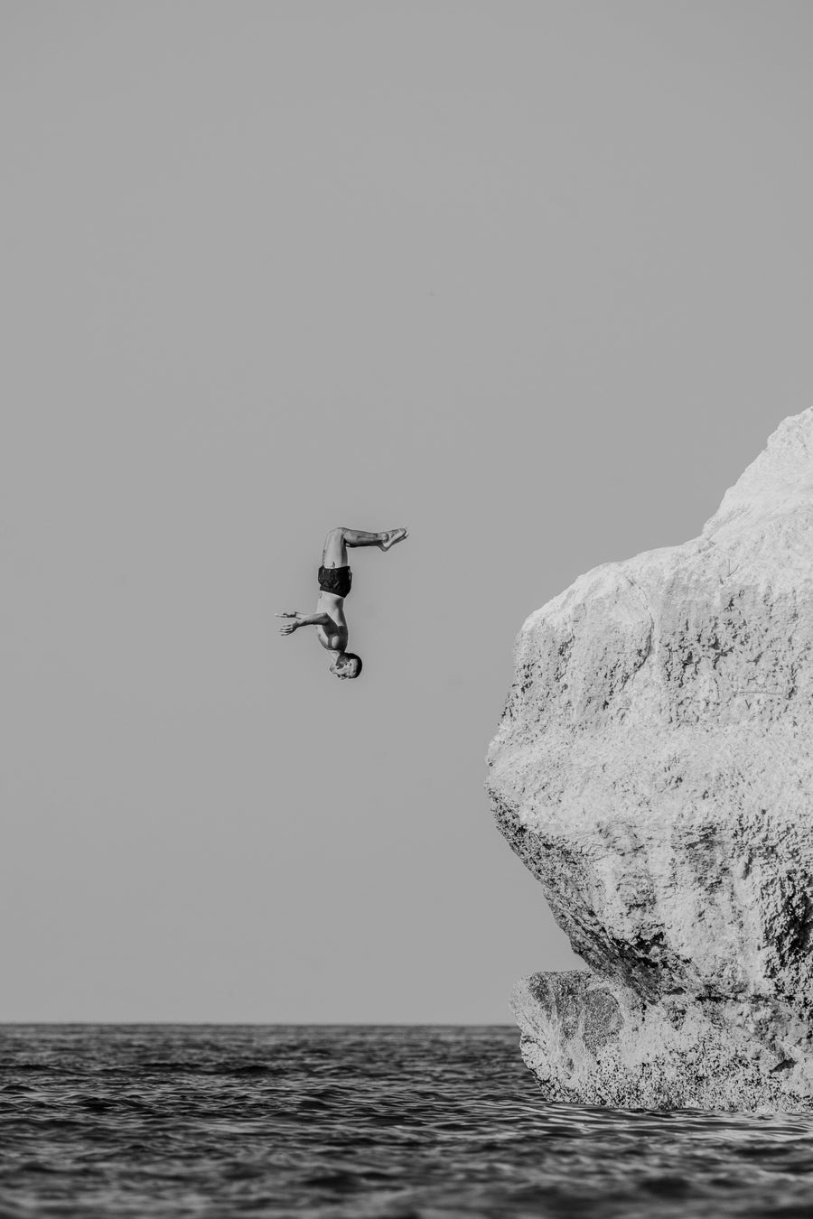 A minimalist black and white photograph of a man performing a backflip off a large white rock into the sea.