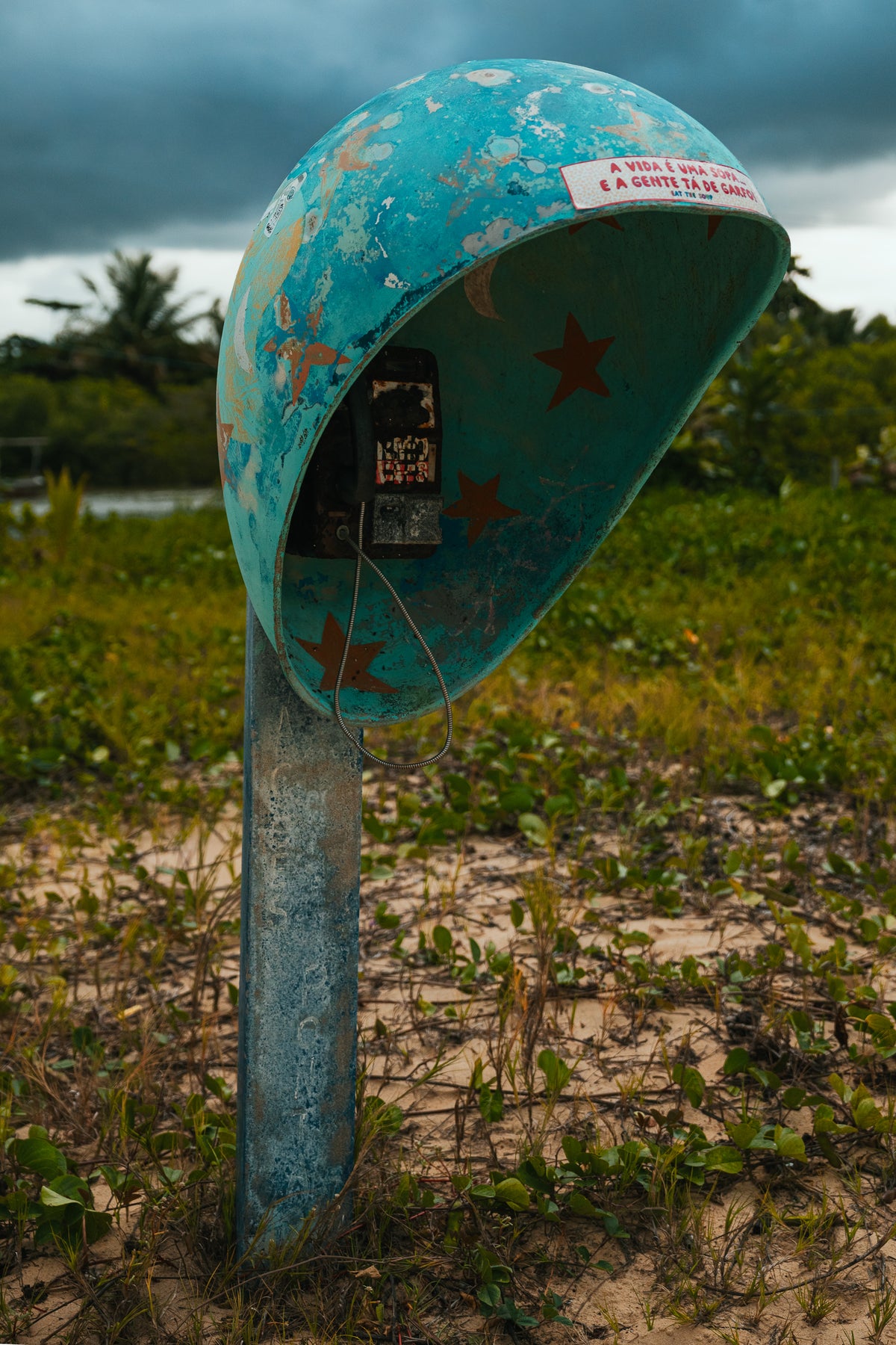 A weathered blue public telephone dome with red stars stands on a sandy beach path in Brazil.
