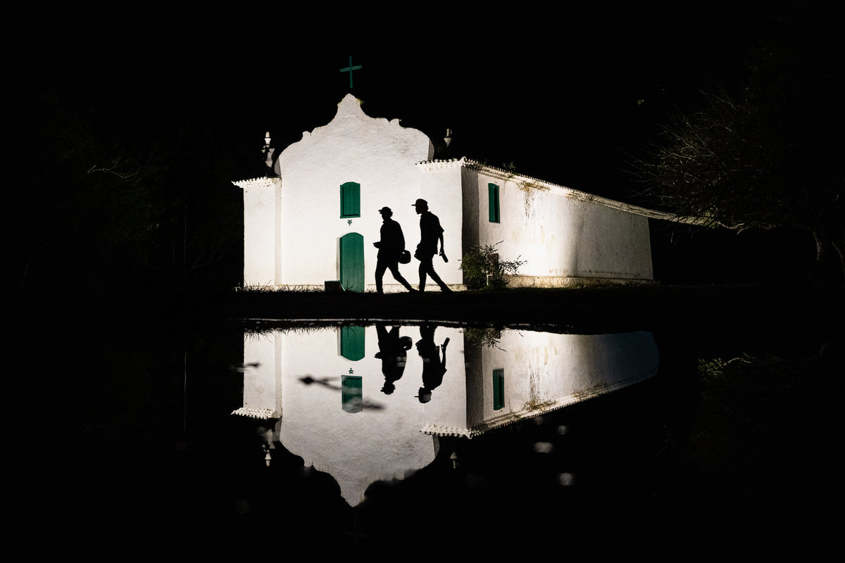 A white colonial church illuminated at night with two silhouetted figures walking past and a clear reflection in a pool of water.