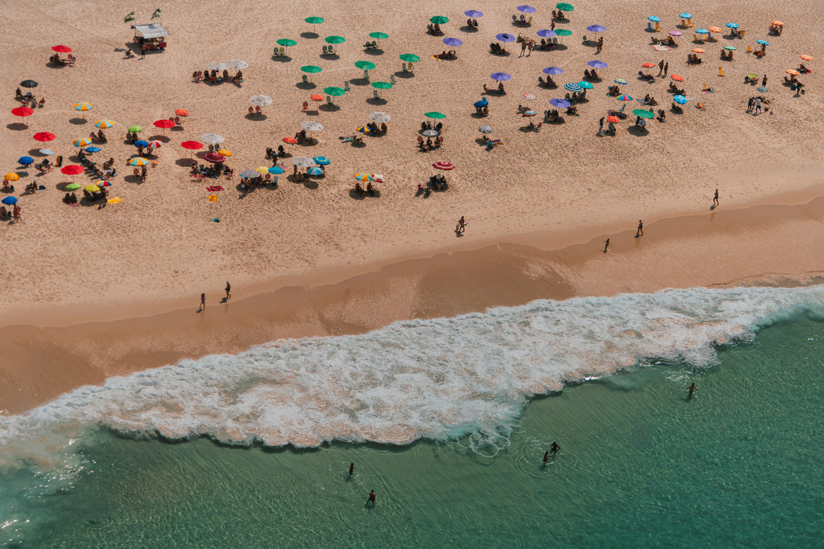High angle aerial view of a sandy beach in Brazil dotted with multi-colored sun umbrellas and people swimming in turquoise water.