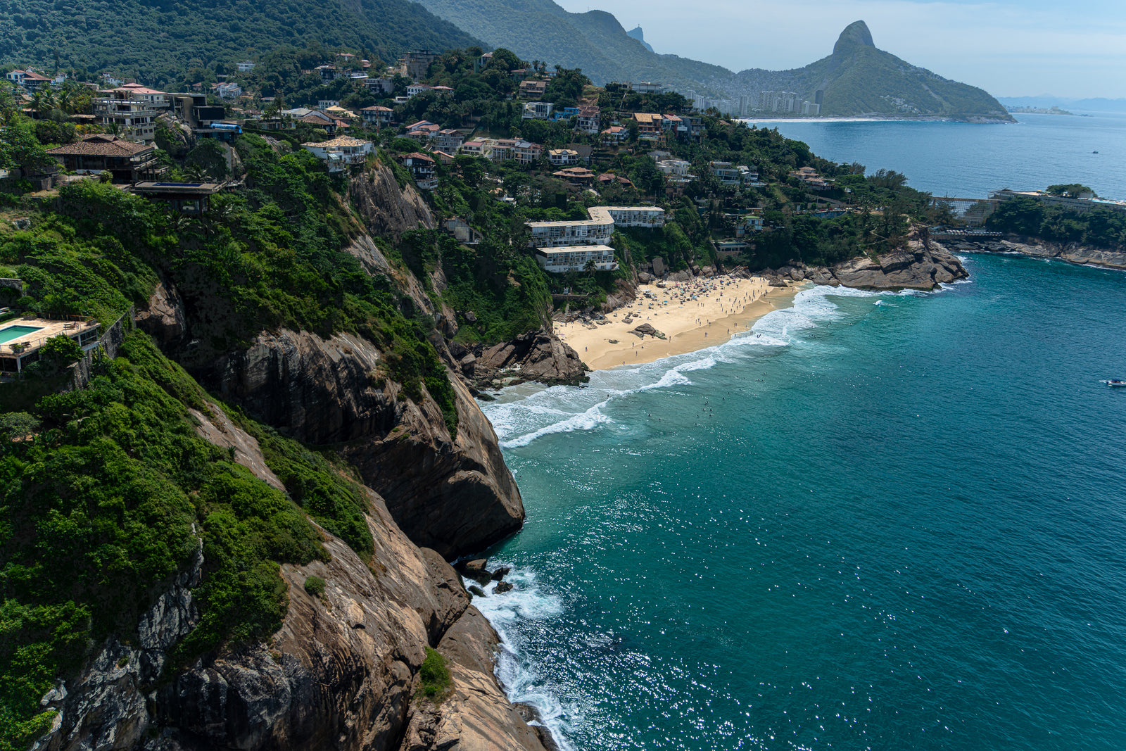 An aerial view of the secluded Praia da Joatinga beach in Rio de Janeiro, Brazil, featuring steep cliffs covered in lush green vegetation and luxury homes overlooking turquoise ocean waves.