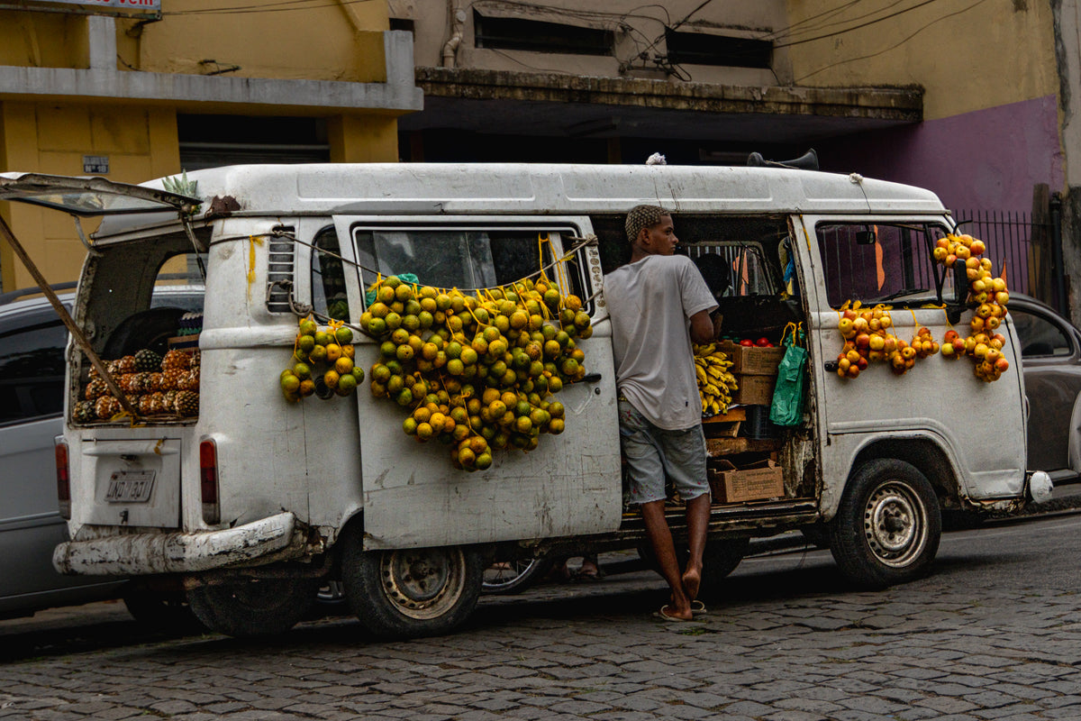 A white vintage Volkswagen Kombi van parked on a cobblestone street in Brazil, decorated with hanging bunches of oranges and pineapples.