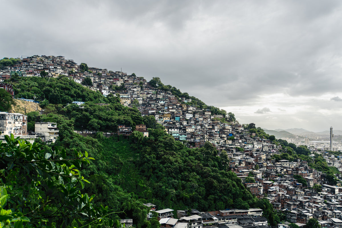 A wide view of a dense Brazilian favela built into a steep green hillside under a cloudy sky.