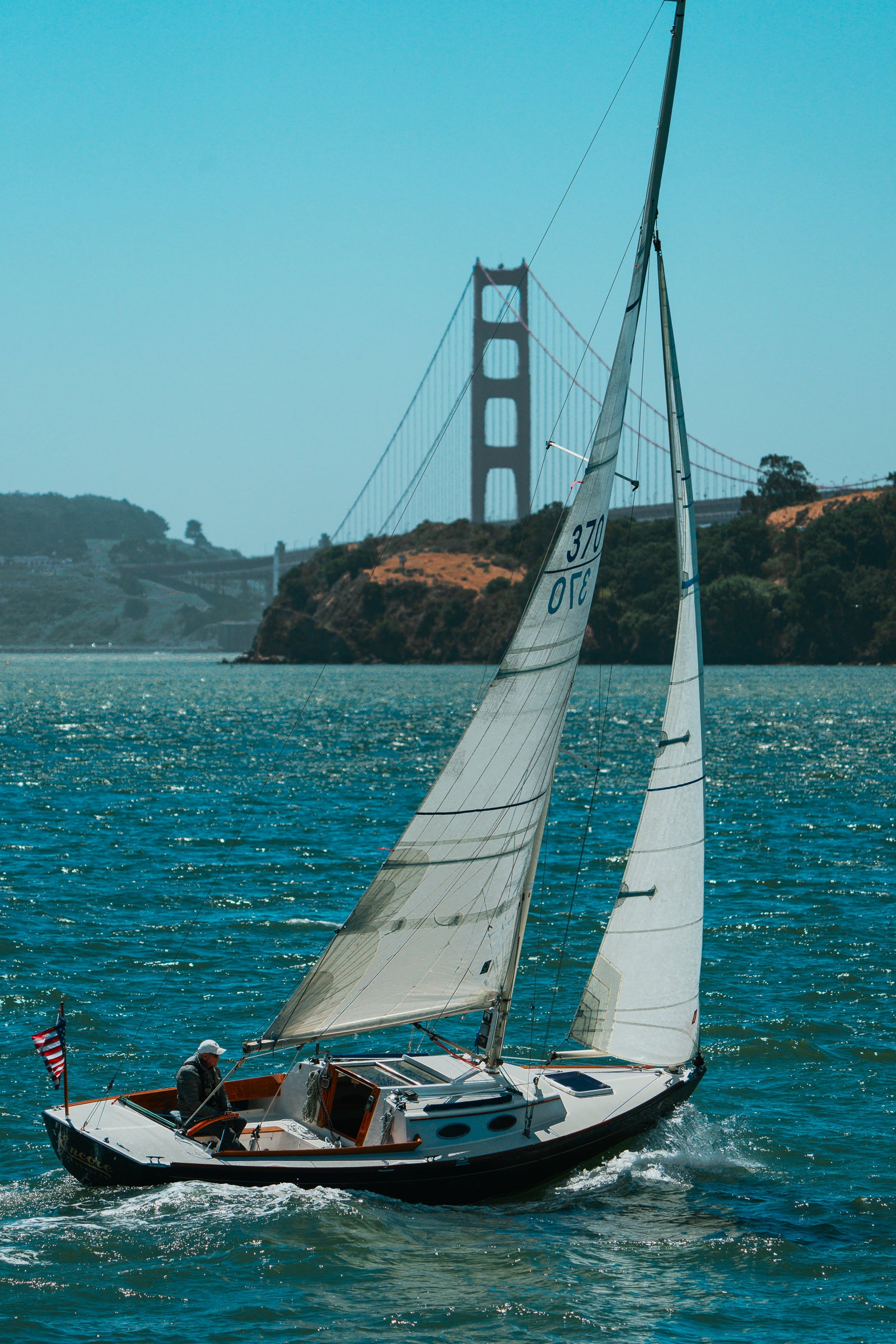 A white sailboat glides across the blue waters of San Francisco Bay with the Golden Gate Bridge visible in the hazy background.
