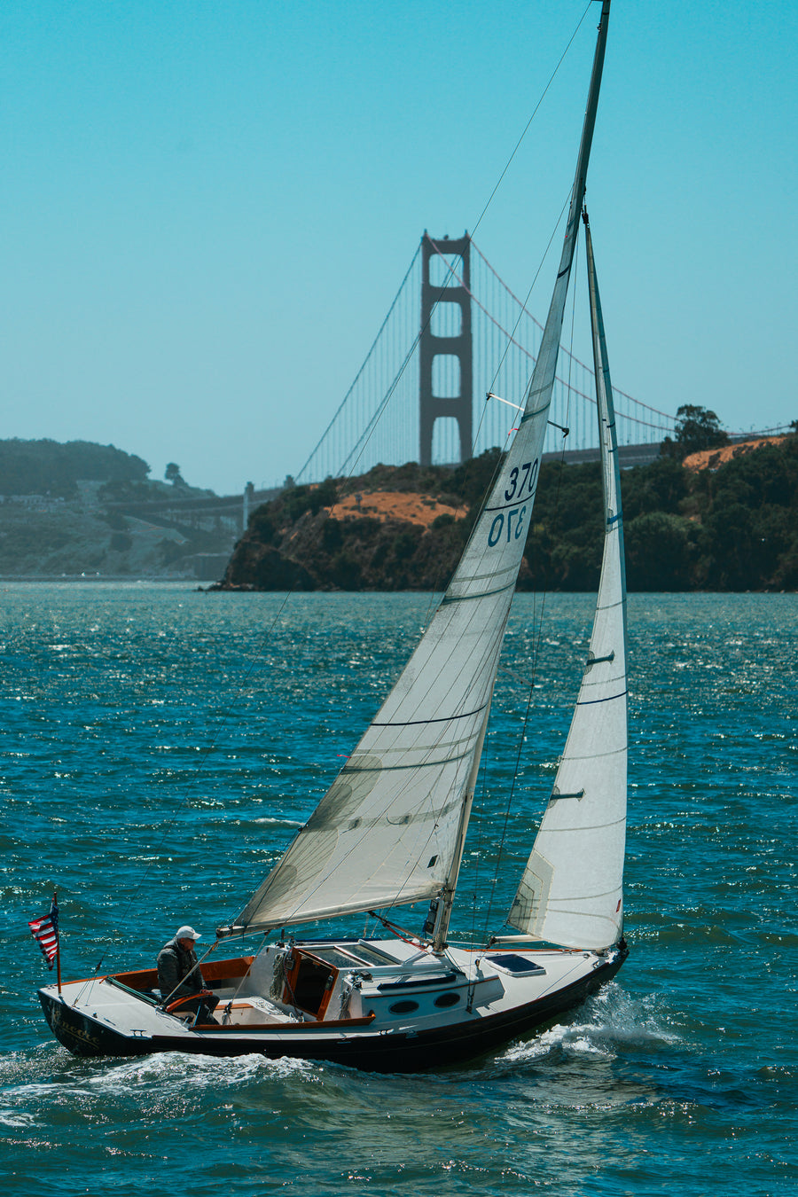 A white sailboat glides across the blue waters of San Francisco Bay with the Golden Gate Bridge visible in the hazy background.