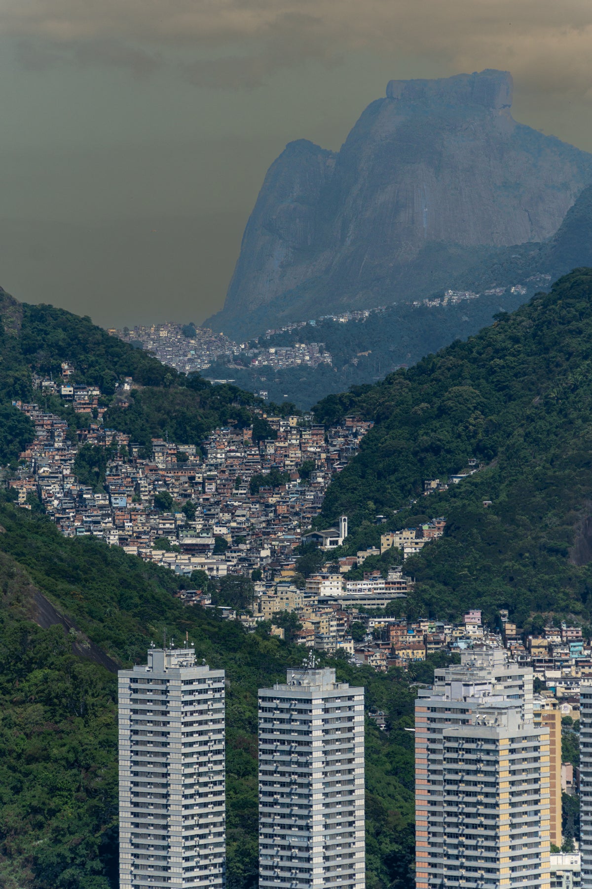 A vertical view of Rio de Janeiro showing modern high-rises in the foreground, a dense hillside favela in the valley, and the massive peak of Pedra da Gávea rising into a misty sky.
