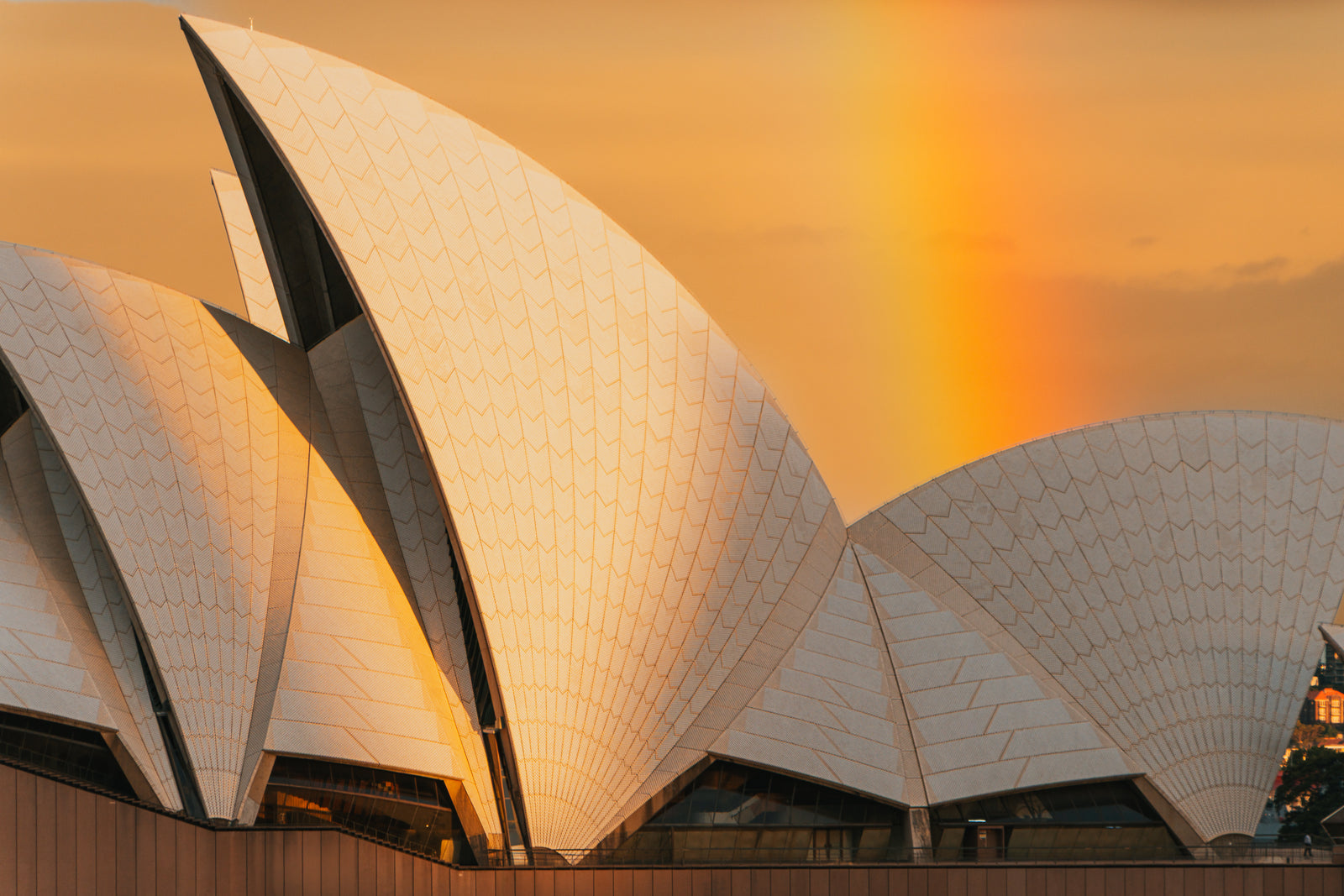 The white tiled sails of the Sydney Opera House are illuminated by a warm sunset with a rainbow in the background.