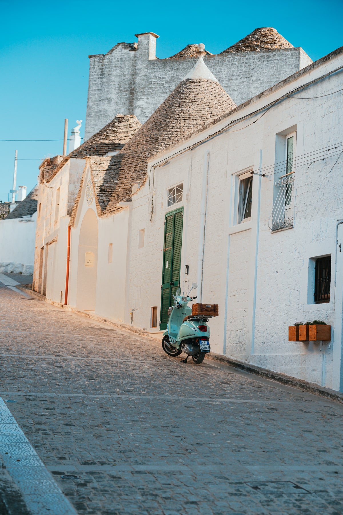 A mint green vintage Vespa scooter parked on a cobblestone street in front of traditional white Trulli houses with conical stone roofs in Italy.