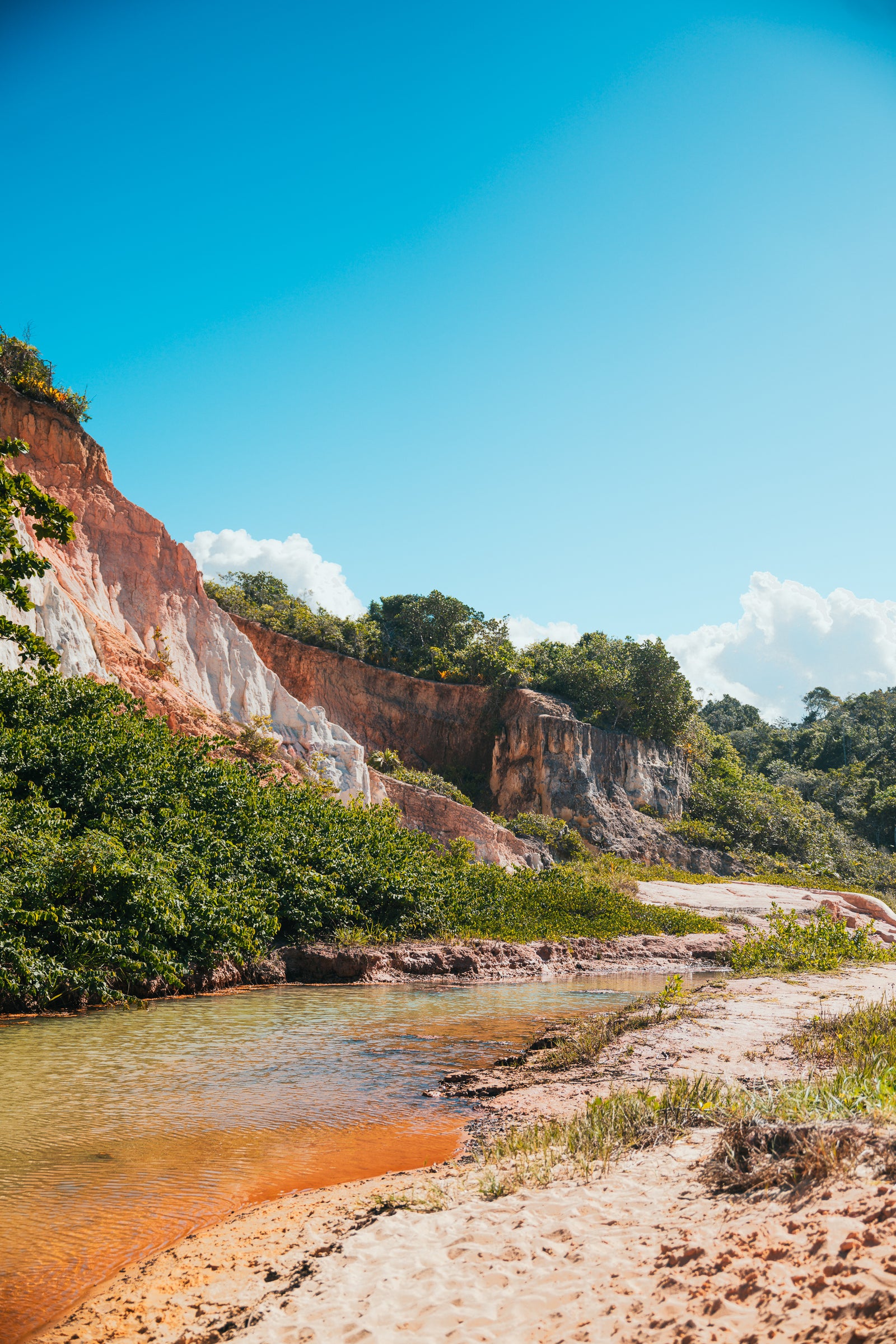 A vertical landscape of red and white clay cliffs bordering a shallow river and sandy beach under a bright blue sky in Brazil.